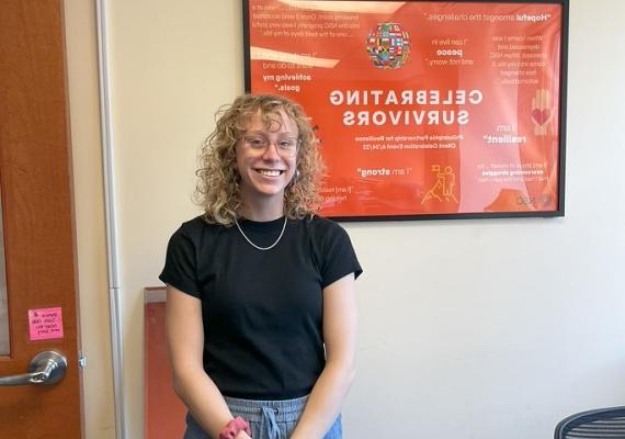 Emma Gross '25 stands in front of a poster at her internship that reads "Celebrating Survivors"